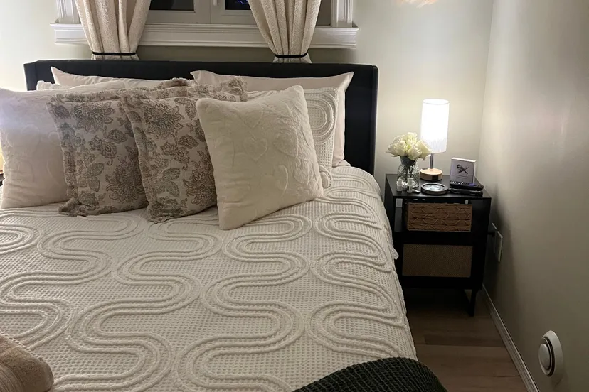 The Aspen Room: a queen bed with textured white bedding, floral pillows and a dark green throw, beside a pale curtained window, pampas wreath and bedside lamp.