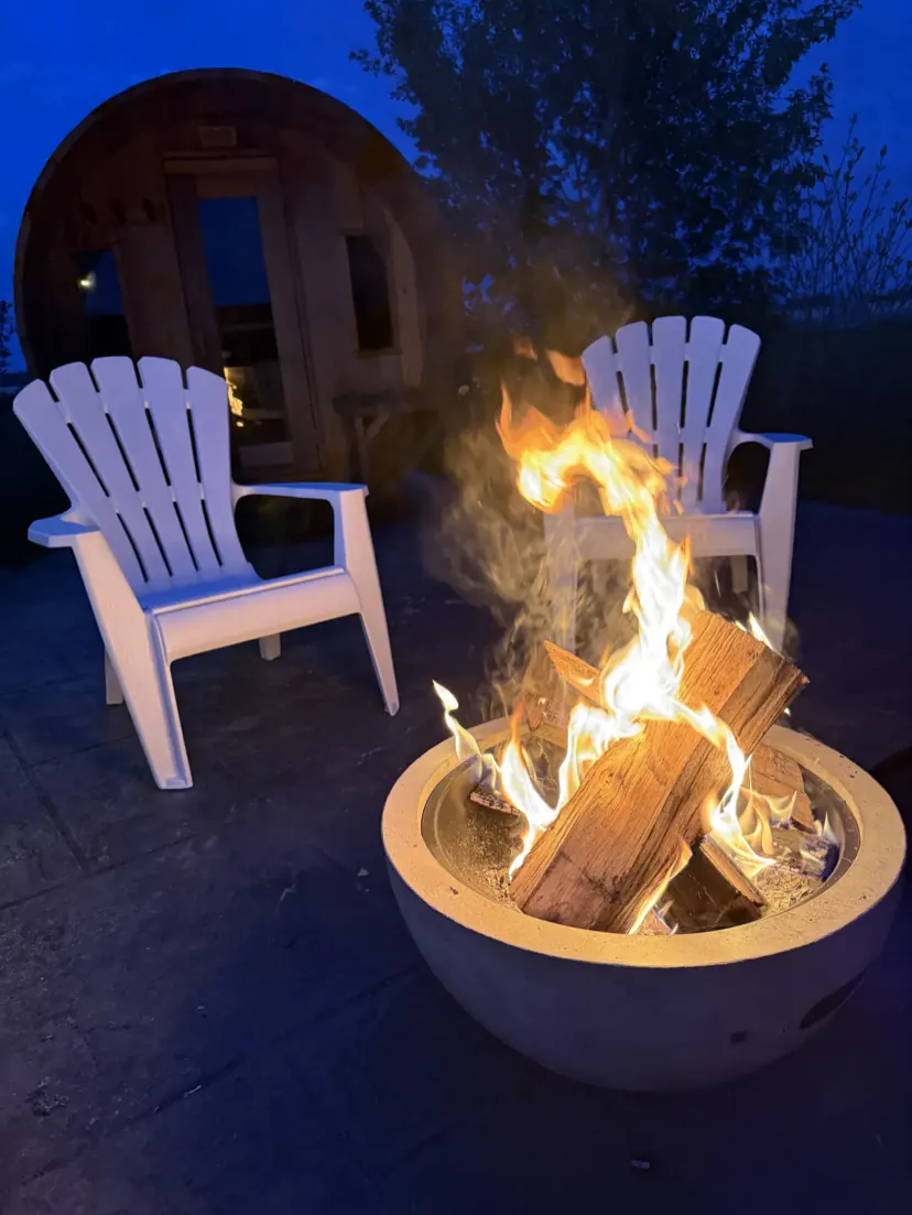 A fire burning in the concrete fire bowl at blue hour, with two white adirondack chairs and the sauna door behind.