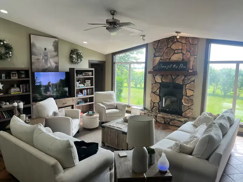 The living room: white sofas and armchairs gathered around a stone fireplace, with bookshelves, a television and tall windows overlooking the lawn.