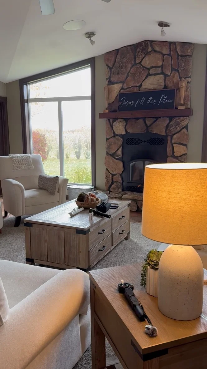 Another view of the living room, with a stone fireplace, cream armchairs, a wooden trunk coffee table and a glowing table lamp.