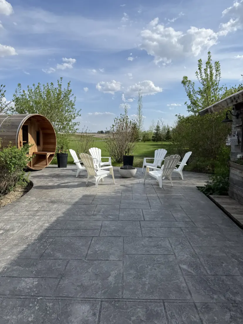 The wide patio with the cedar barrel sauna and a circle of white adirondack chairs, with lawn, trees and a big sky beyond.