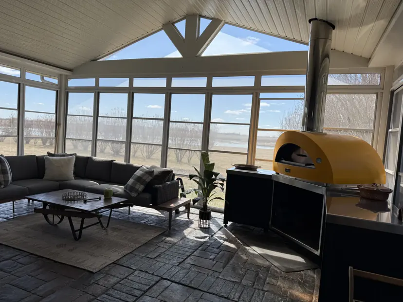 The sunroom: a vaulted white ceiling, windows on three sides looking out to the prairie, a dark sectional sofa and a yellow wood-fired pizza oven.